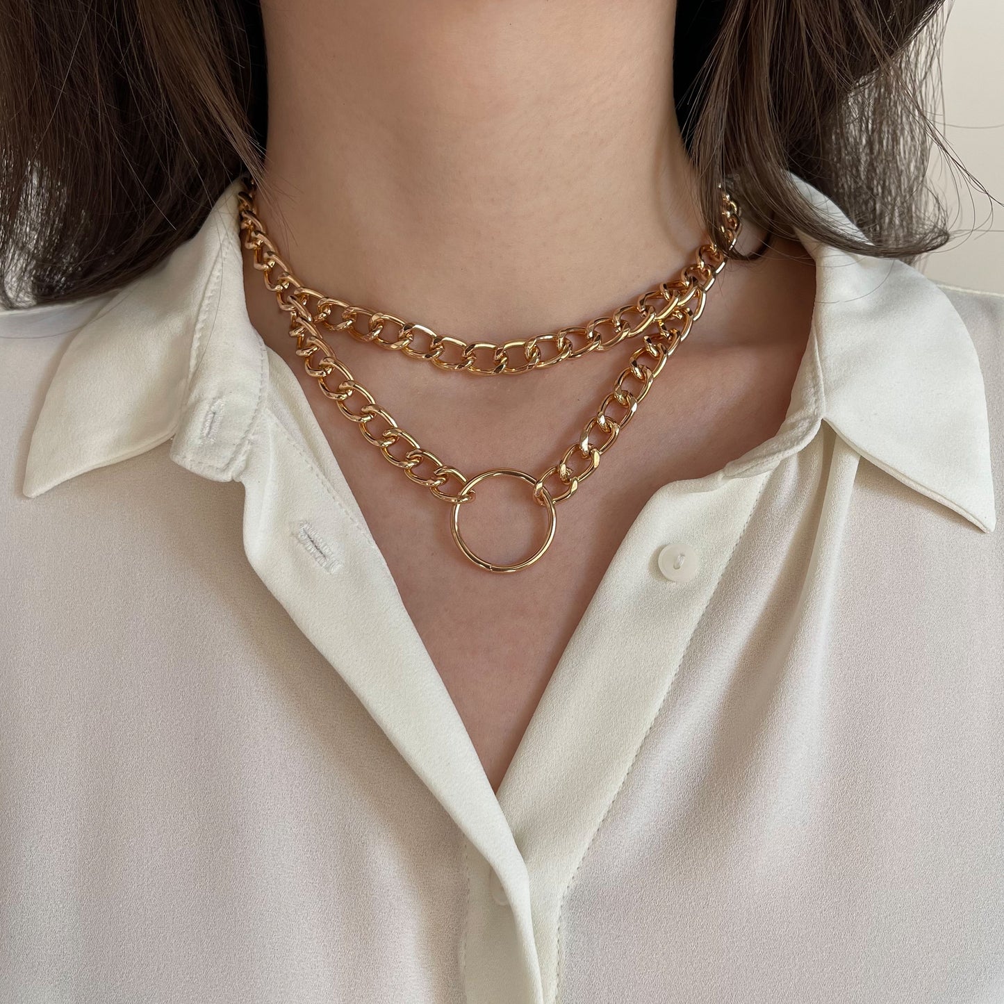 Chunky double gold chain choker featuring a round centerpiece ring. Made from polished metal, the necklace rests on a woman’s neck over a white blouse. The close-up frontal shot emphasizes its structured drape and contemporary style for modern fashion looks.