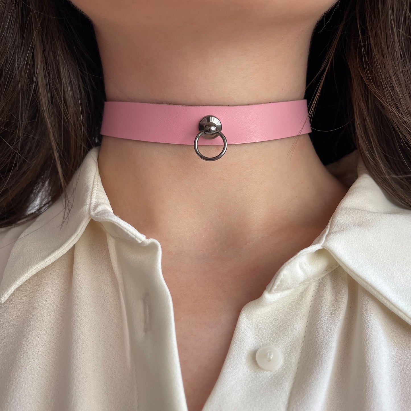 Minimalist pink leather choker with central dark silver-tone ring and rivet. Made from semi-matte genuine leather, 1.5 cm (0.59 in) wide. Shown on a woman’s neck in a white shirt, adding a delicate, romantic highlight to the neckline.