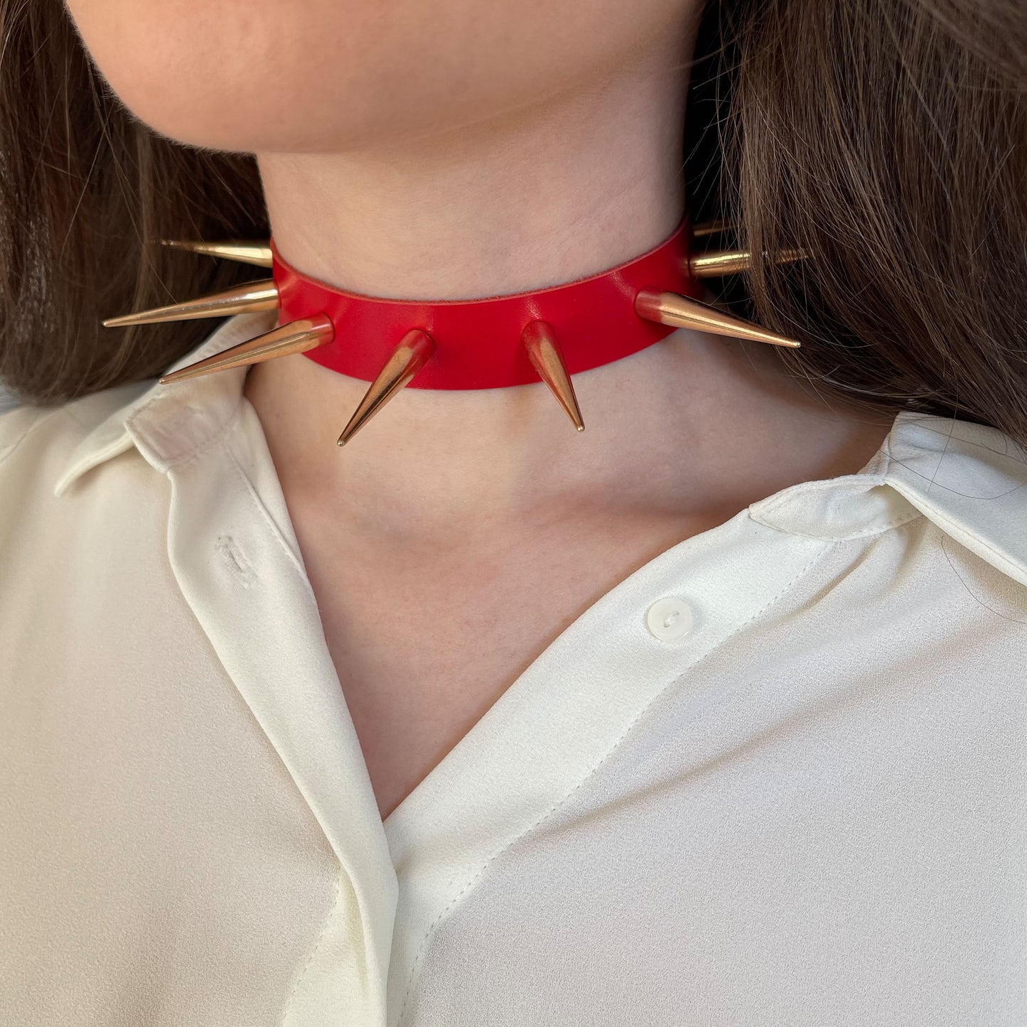 Bright red leather choker with long gold-tone metal spikes, shown from the side on a woman's neck wearing a white shirt. The sharp cone studs radiate a bold punk-inspired aesthetic. The choker is crafted from semi-matte genuine leather and fastened securely at the back.