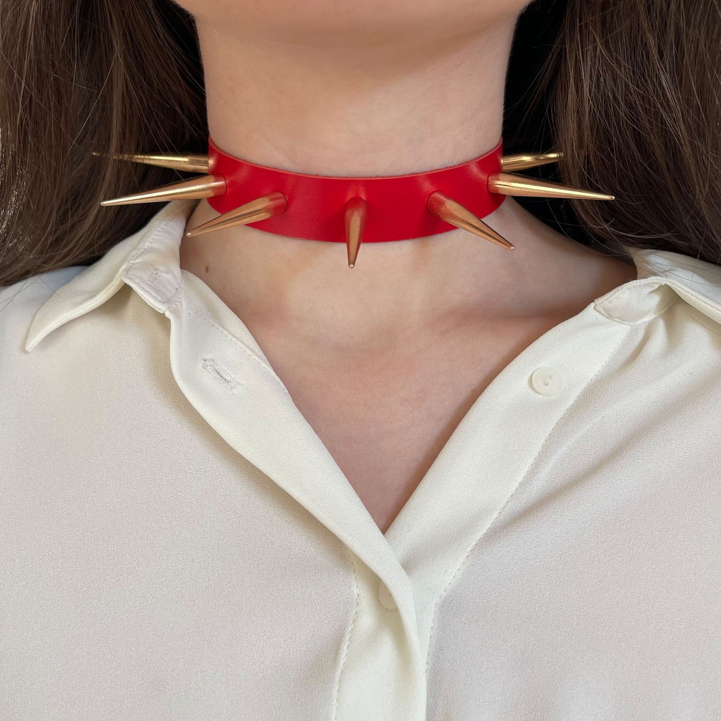 Front view of a red leather choker featuring oversized gold-tone spikes evenly spaced around the neck. Made from genuine leather with a semi-matte finish, this statement accessory contrasts dramatically with a light blouse and channels rebellious punk energy.