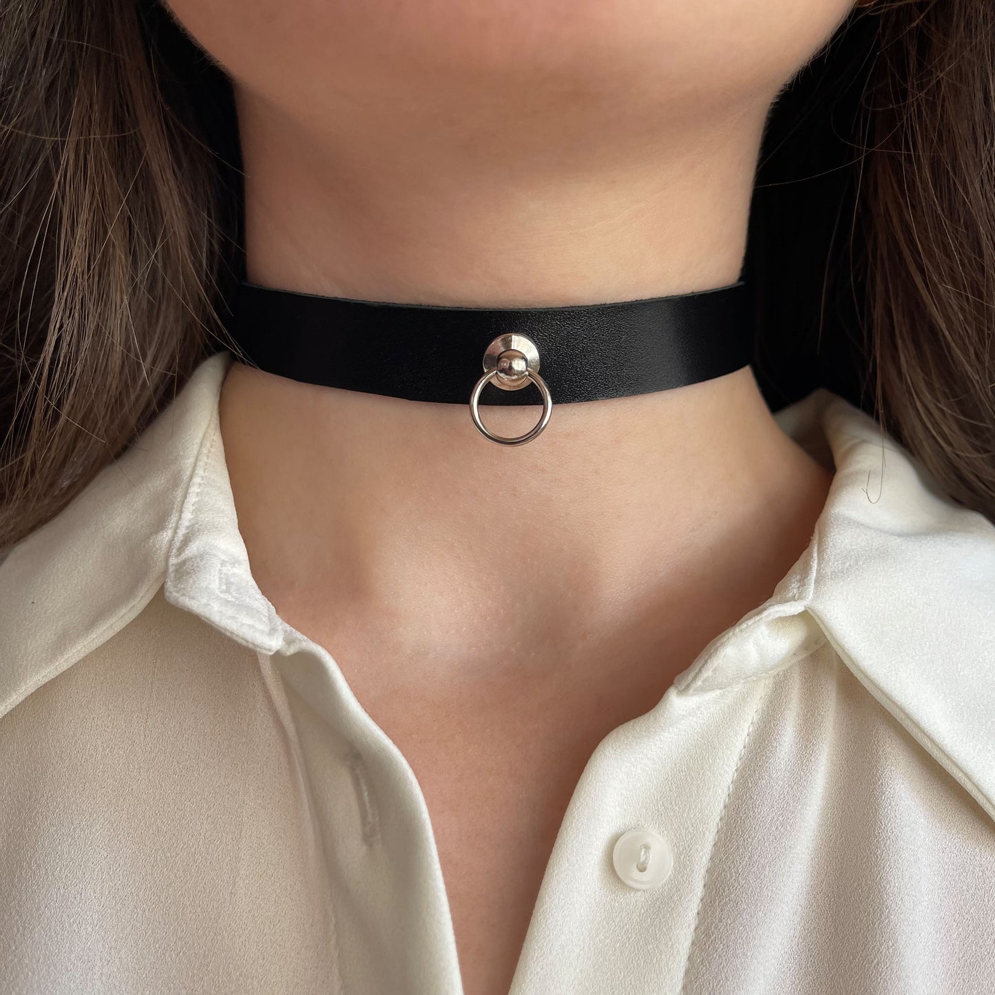Classic black leather choker with a silver ring detail at the center. Crafted from genuine semi-matte leather, 1.5 cm (0.59 in) wide and 1.4–1.8 mm thick. Buckle closure. Close-up of a woman’s neck in a white shirt, highlighting the contrast and bold simplicity of the piece.