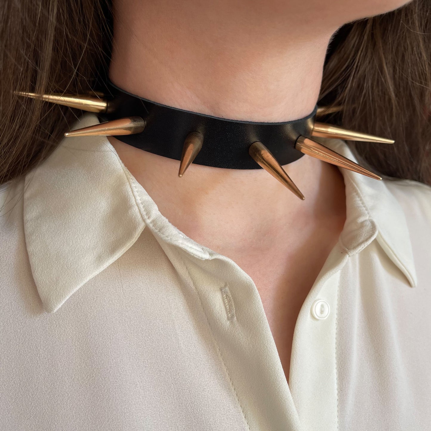 Minimalist black leather choker with large, glossy gold-tone metal spikes. Side view highlights the aggressive yet elegant design, worn over a white blouse. The spikes form a symmetrical halo around the neck, creating a strong, fashion-forward look.
