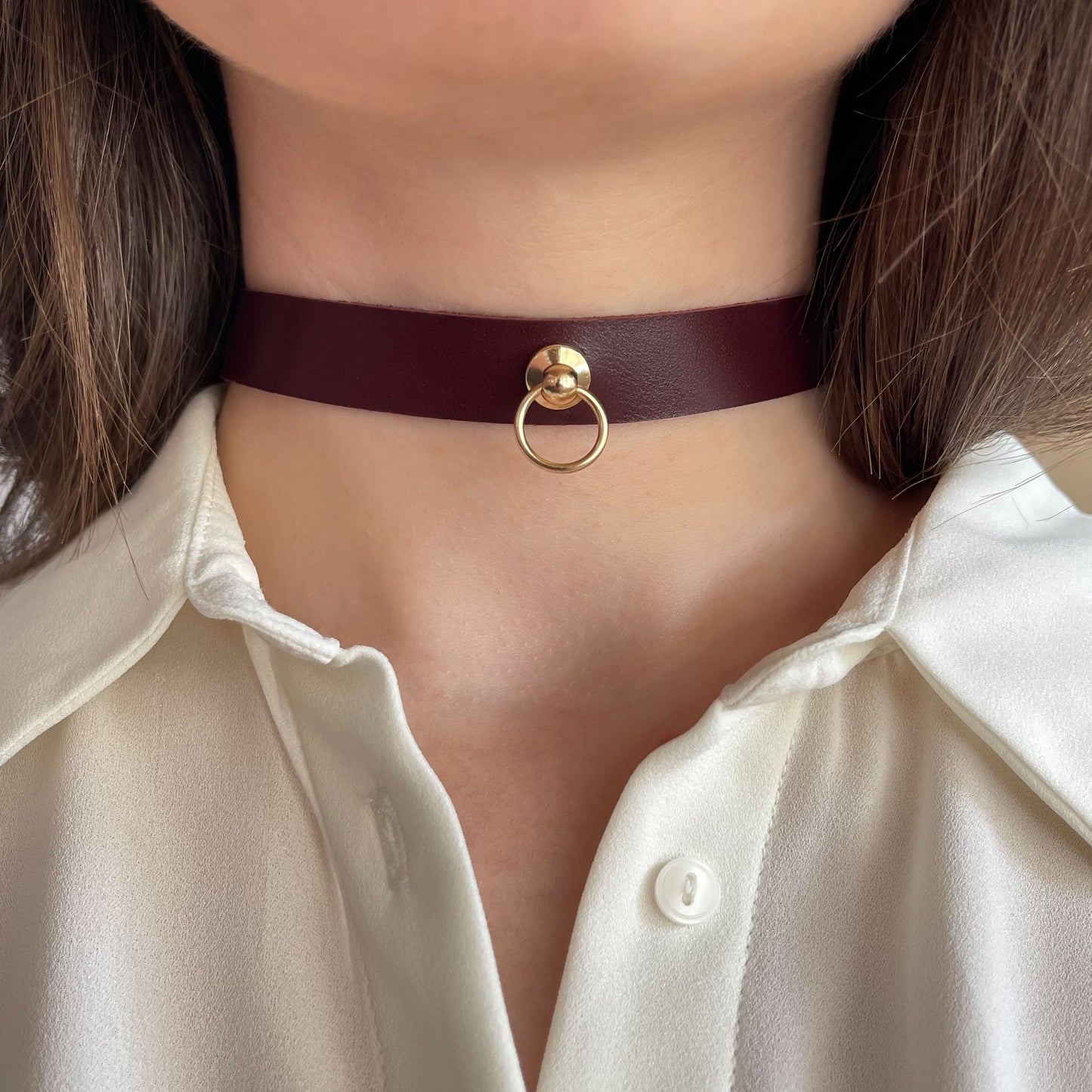 Minimalist marsala choker with a gold-tone ring and rivet. Genuine semi-matte leather, 1.5 cm (0.59 in) wide, 1.4–1.8 mm thick. Shown front-facing on a woman's neck in a white shirt, this accessory blends elegance with a subtle provocative edge.