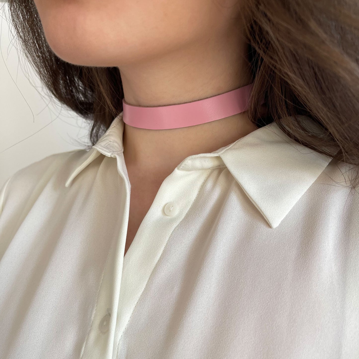 Soft pink leather choker in minimalist style, 1.5 cm (0.59 in) wide, made from semi-matte genuine leather (1.4–1.8 mm thick), with a classic buckle. Shown on a woman in a white blouse, the pastel tone adds a gentle feminine accent. A light, elegant piece for casual and romantic outfits.