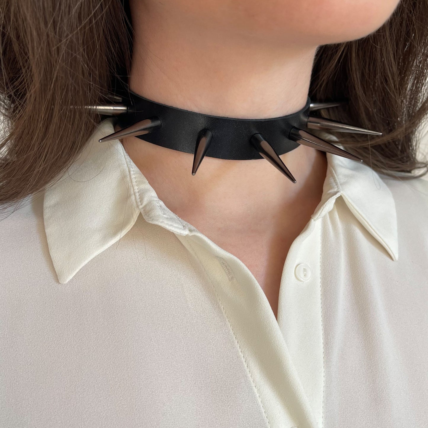 Side view of a black leather choker featuring glossy black cone spikes. Worn on a woman’s neck with a soft white blouse, this accessory captures bold contrast and gothic minimalism in a clean, streamlined form.