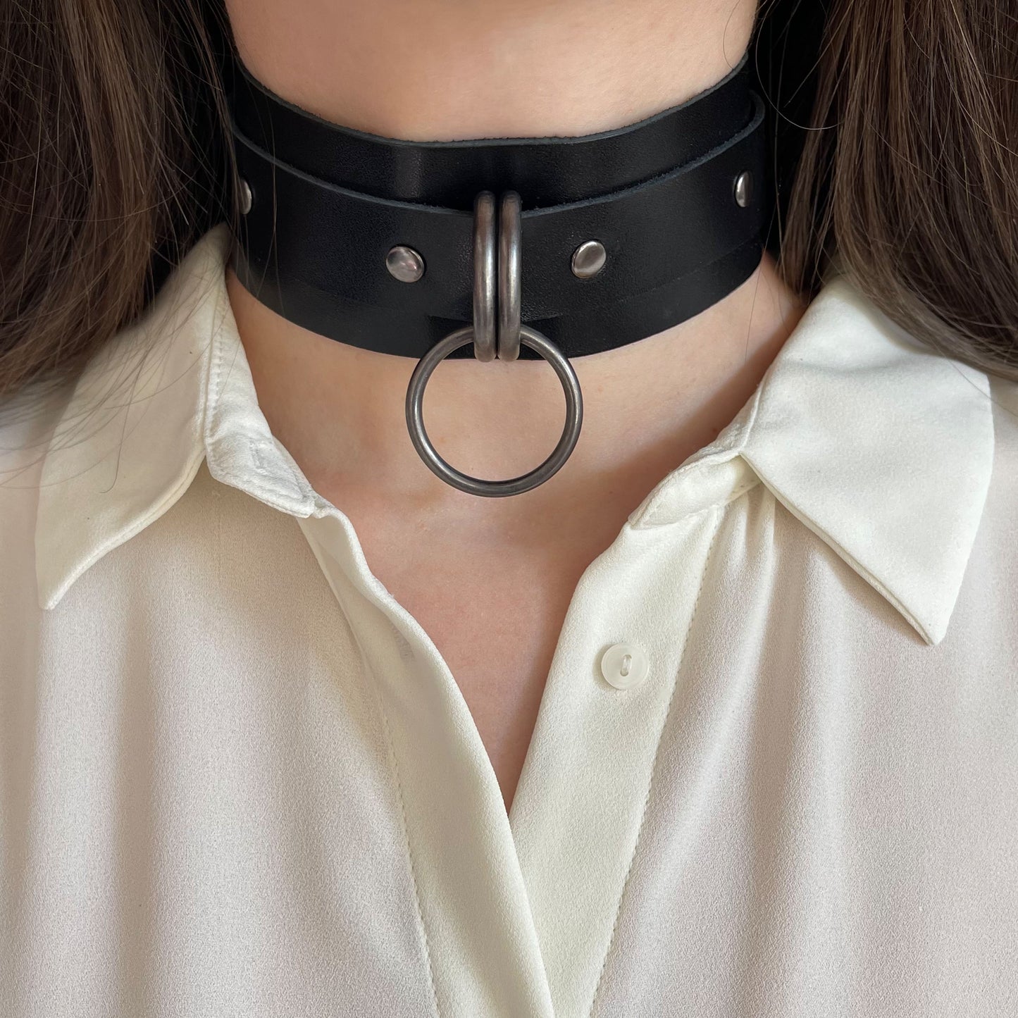 Front view of a wide black leather choker with dark nickel hardware worn on the neck. The round 25 mm O-ring is centered between two D-rings on the wide leather band. The straight cut leather edges and solid metal elements are visible.
