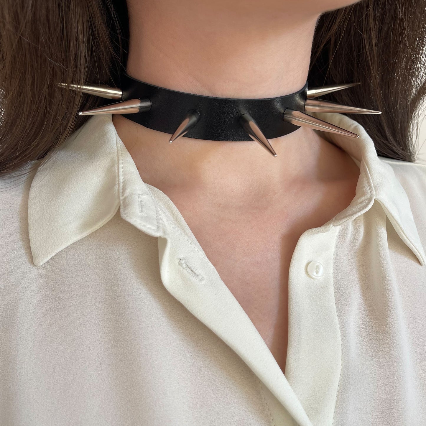 Wide black leather choker adorned with large polished silver-tone metal spikes. Side view on a woman's neck over a cream blouse. This statement collar combines classic punk details with refined, modern execution.