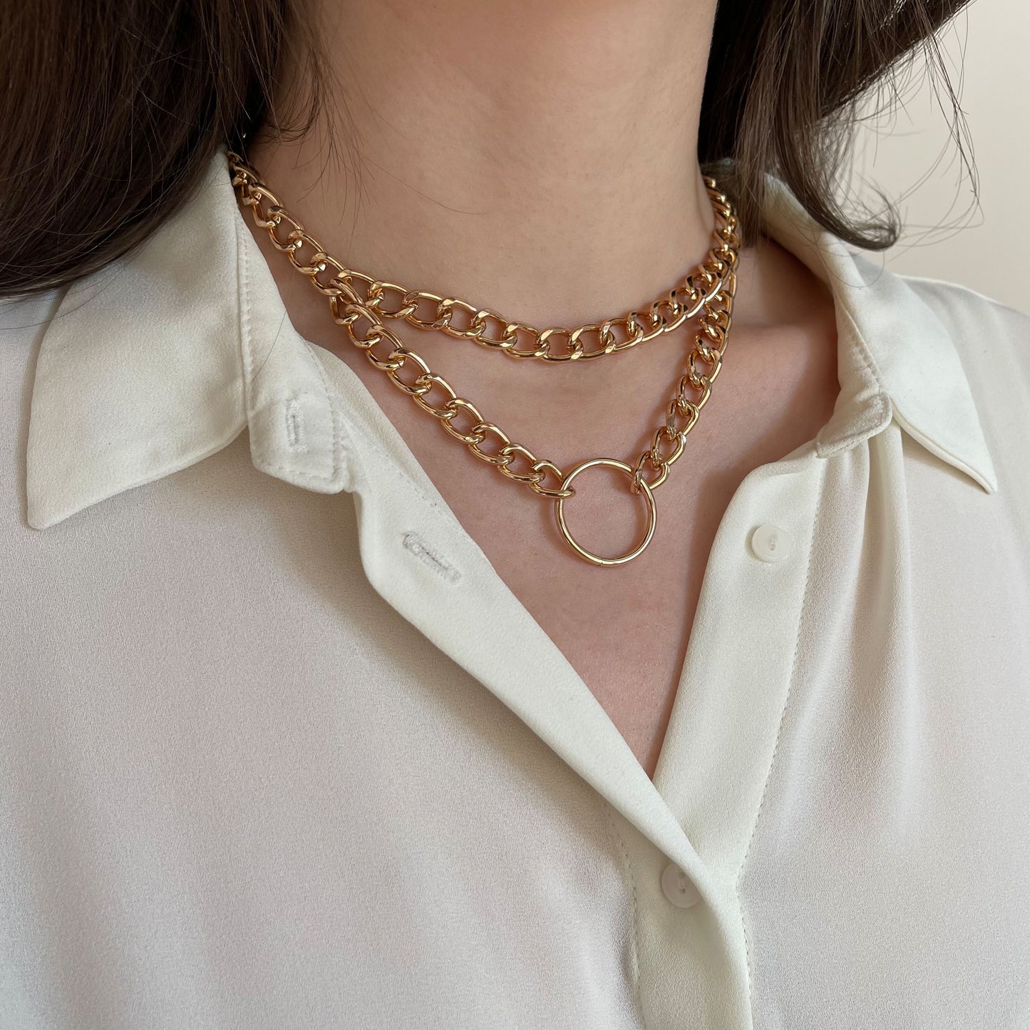 Double-layer gold-tone chain choker with a central metal ring. Worn on a woman’s neck over a white shirt, side angle shows the glossy finish and bold statement look. Lightweight and adjustable, this collar-style necklace blends minimalism with a touch of edgy elegance.