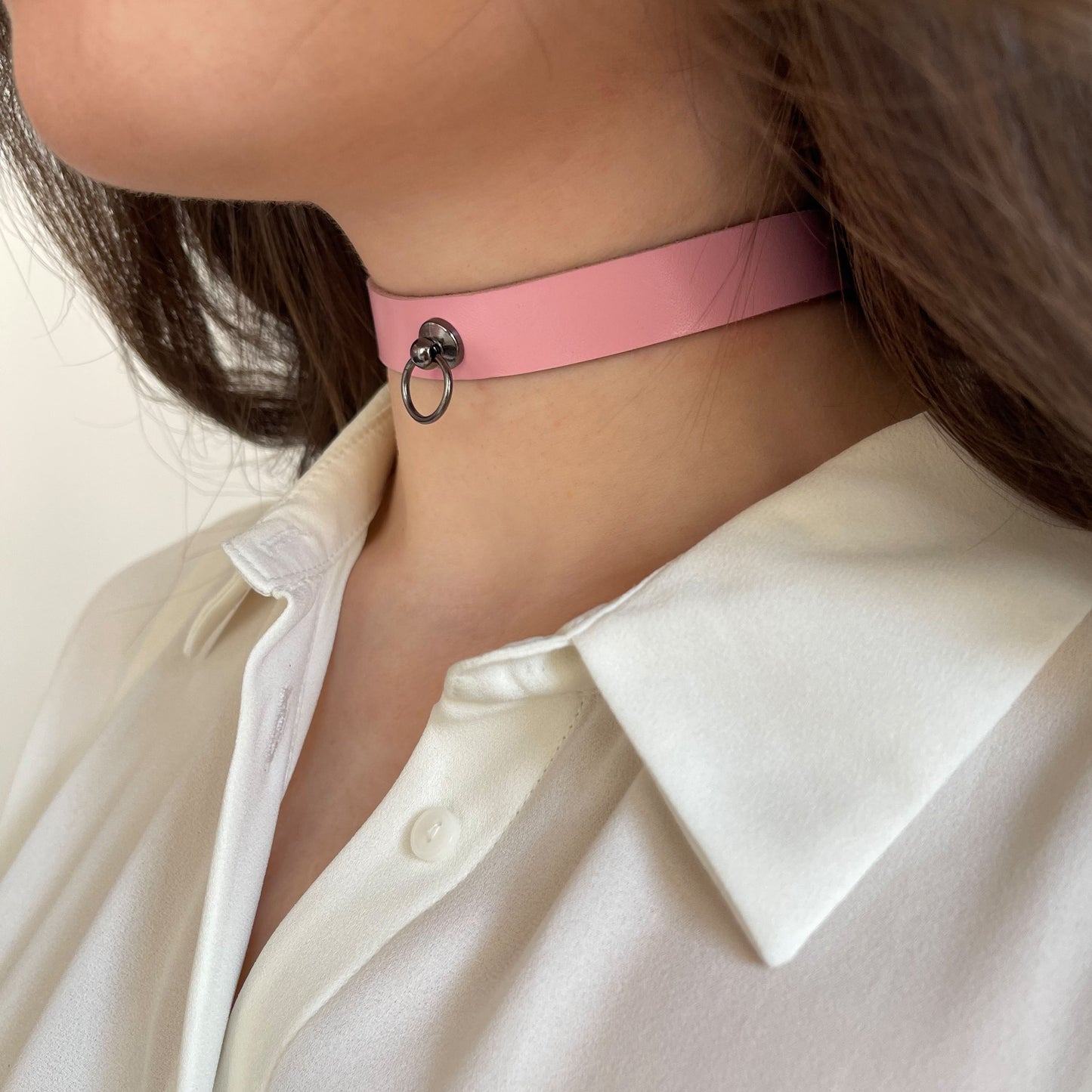 Soft pink leather choker with a dark metal ring in the center. Made of genuine semi-matte leather, 1.5 cm (0.59 in) wide, 1.4–1.8 mm thick. Side profile of a woman’s neck in a light blouse, accentuating the pastel tone and feminine character.