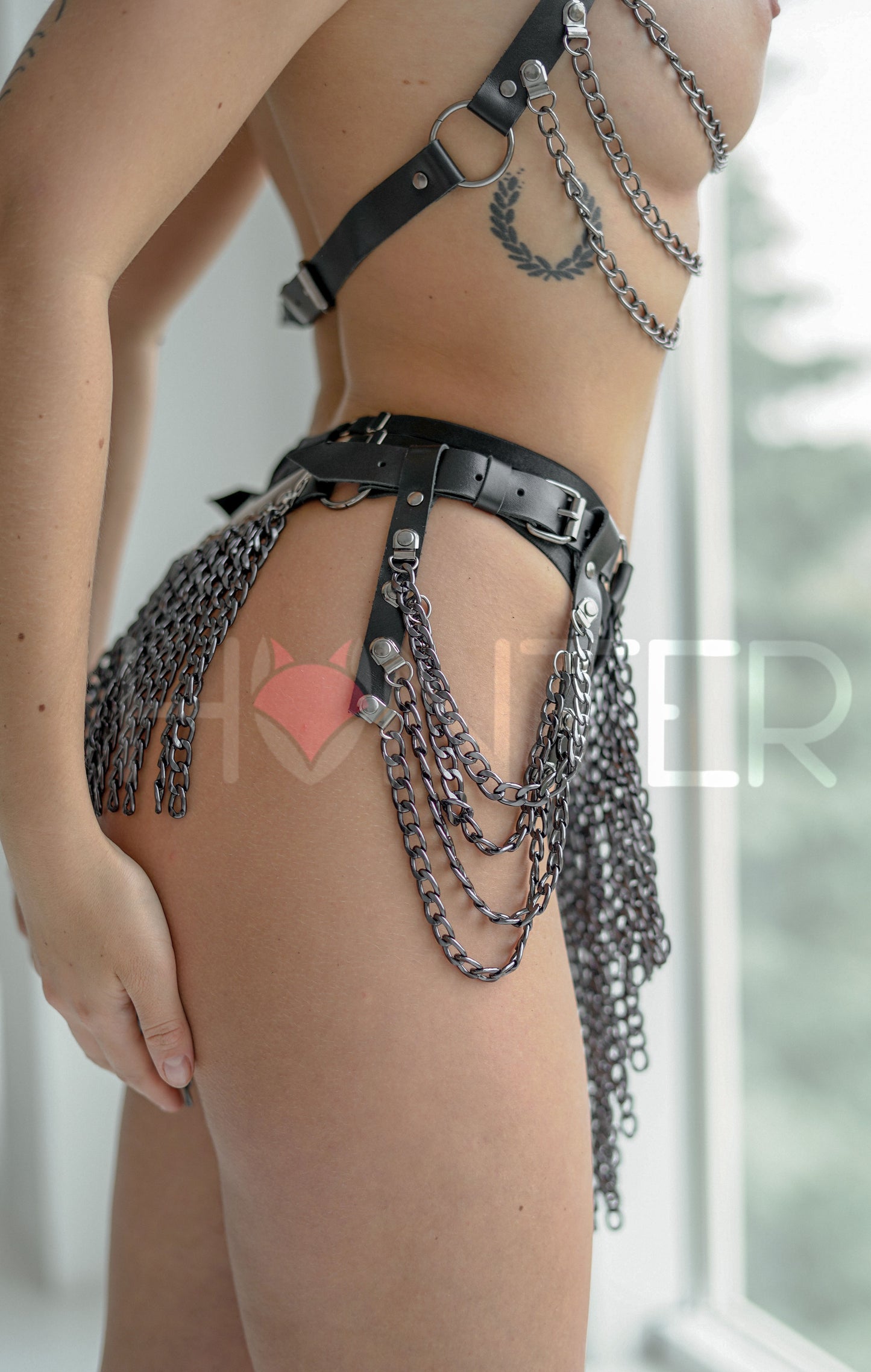 Close side detail of the chain drape chest harness in genuine black leather with dark nickel hardware, showing the side ring, the buckle strap under the arm, and several curved chain rows attached along the leather edge of the chest piece.