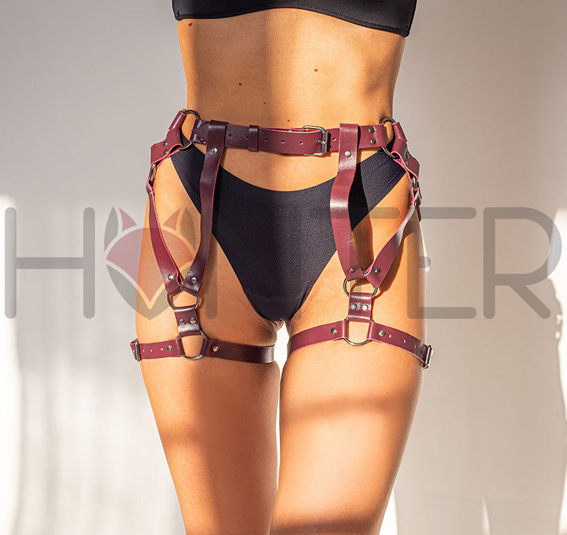 Front view of a model wearing a marsala leather garter belt with dark nickel hardware over black underwear. The photo shows the front waist strap, vertical leather sections, mixed ring sizes, geometric strap layout on both thighs, and lower thigh straps fastened around the legs.