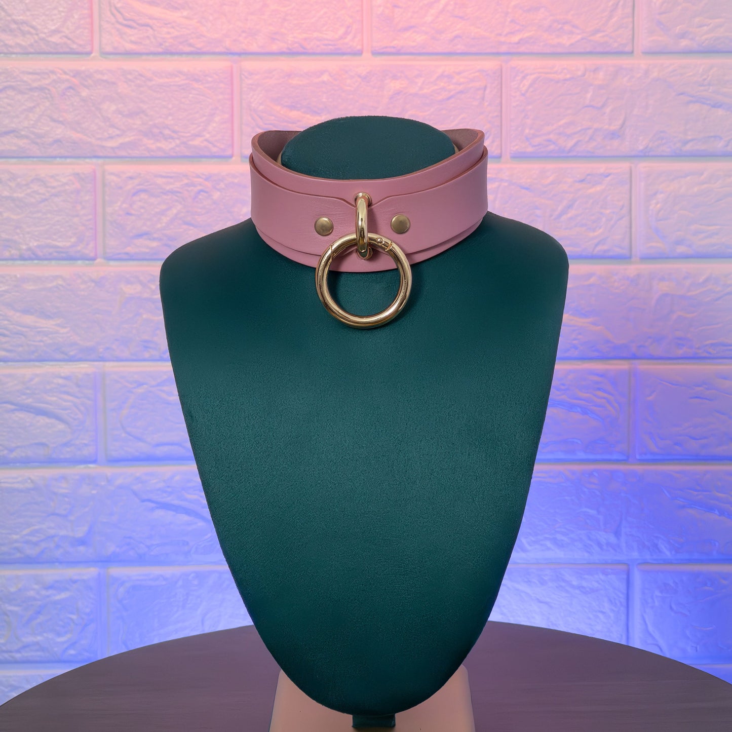 Pink leather choker with gold ring on a green mannequin against a textured brick wall.
