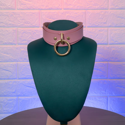 Pink leather choker with gold ring on a green mannequin against a textured brick wall.