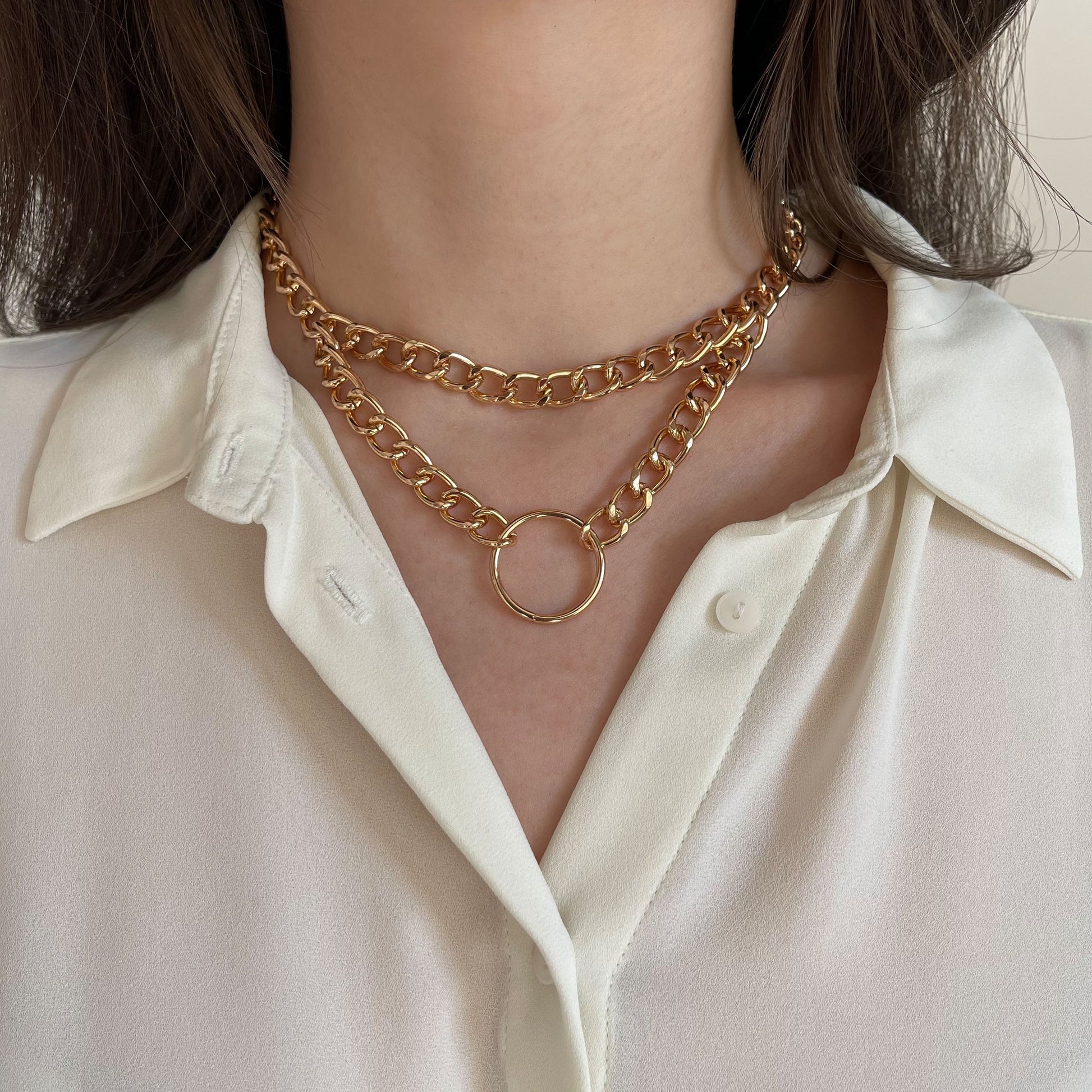 Chunky double gold chain choker featuring a round centerpiece ring. Made from polished metal, the necklace rests on a woman’s neck over a white blouse. The close-up frontal shot emphasizes its structured drape and contemporary style for modern fashion looks.