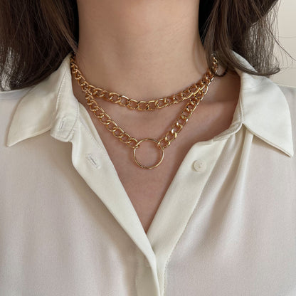 Chunky double gold chain choker featuring a round centerpiece ring. Made from polished metal, the necklace rests on a woman’s neck over a white blouse. The close-up frontal shot emphasizes its structured drape and contemporary style for modern fashion looks.