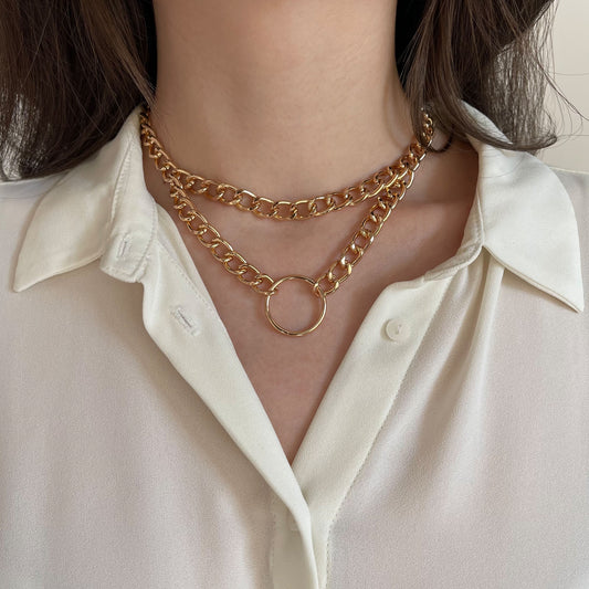 Chunky double gold chain choker featuring a round centerpiece ring. Made from polished metal, the necklace rests on a woman’s neck over a white blouse. The close-up frontal shot emphasizes its structured drape and contemporary style for modern fashion looks.