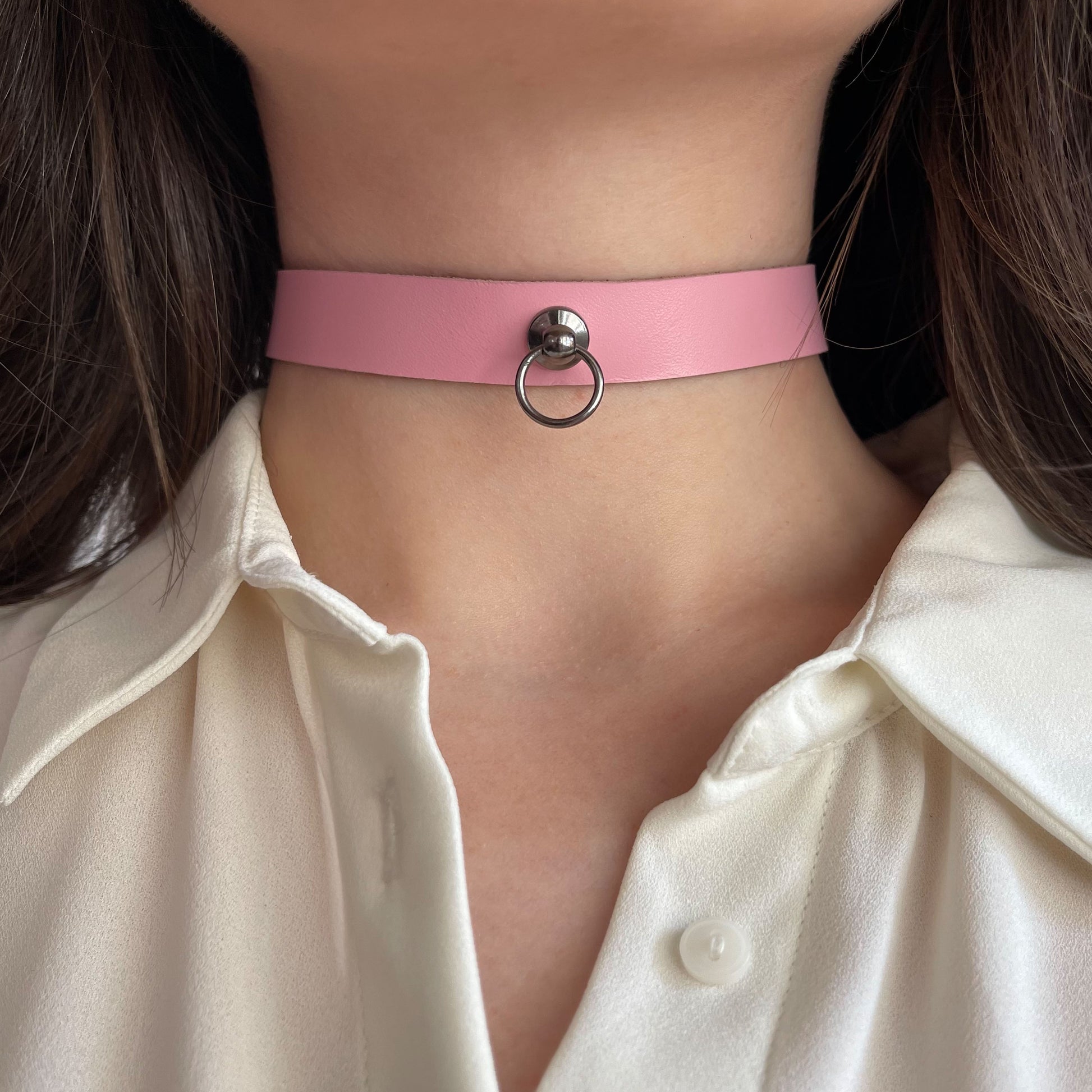 Minimalist pink leather choker with central dark silver-tone ring and rivet. Made from semi-matte genuine leather, 1.5 cm (0.59 in) wide. Shown on a woman’s neck in a white shirt, adding a delicate, romantic highlight to the neckline.