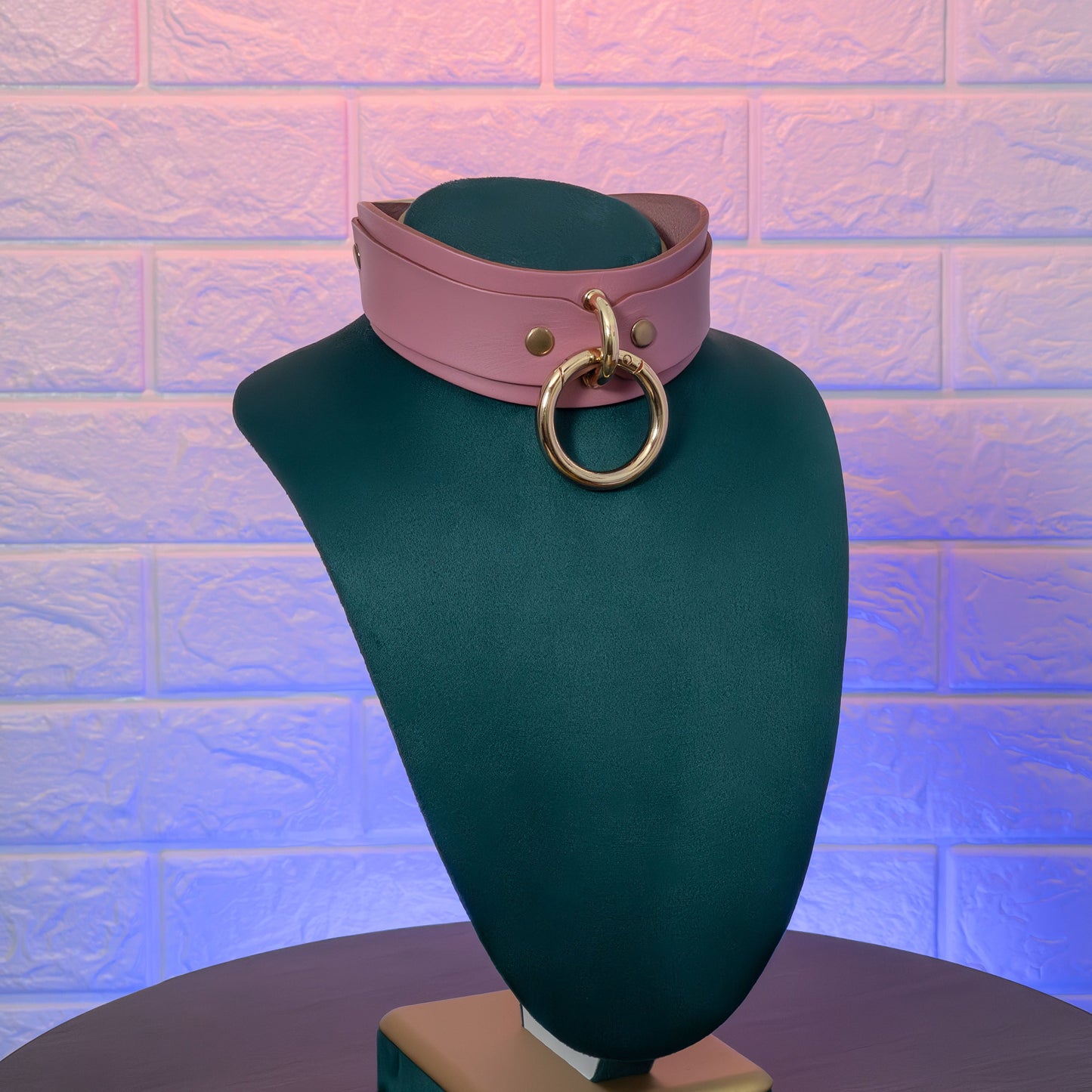 Pink leather choker with gold ring on a green mannequin against a textured brick wall.