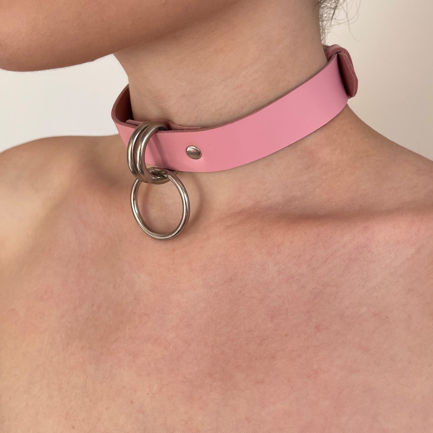 Pink leather choker worn on the neck in a semi-profile view, two silver D-rings at the front holding a round O-ring, smooth 20 mm wide strap with a straight cut edge, metal ring hanging slightly forward from the collar.