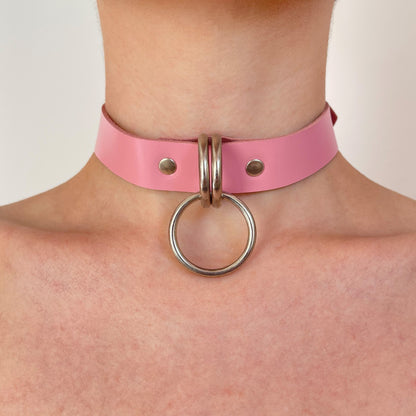 Pink leather choker shown from the front, two D-rings aligned at the center holding a round silver O-ring, leather strap evenly wrapped around the neck, straight cut edge visible, polished metal hardware as the main focus.