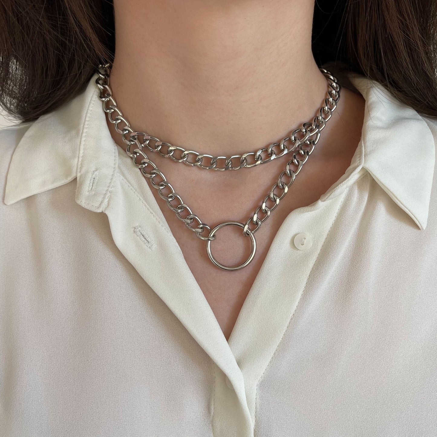 Bold silver double chain choker with a central ring accent. Resting on a woman’s neck over a cream shirt, the polished metal reflects light, creating a clean and futuristic aesthetic. A standout accessory for contemporary or urban styling.