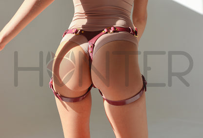 Rear view of a person wearing a flesh-toned bodysuit with a dark red leather harness-style belt and matching leg straps, against a plain light background.
