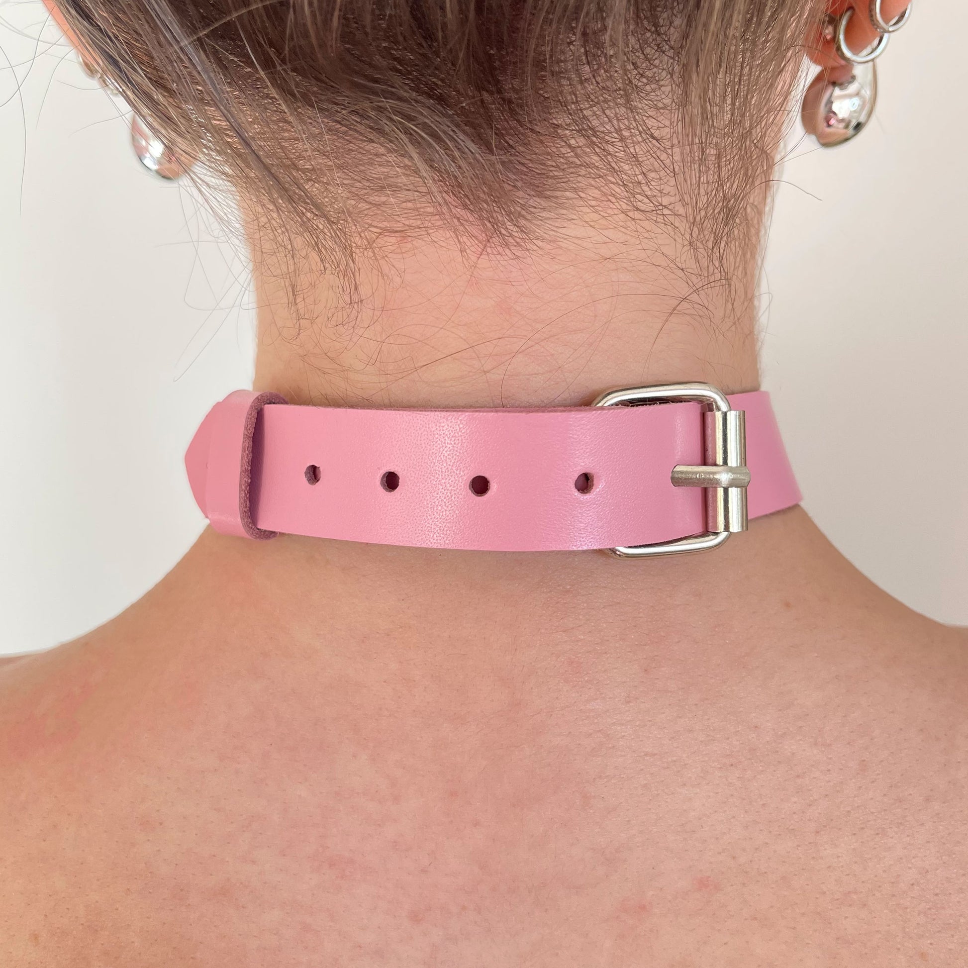 Back view of a pink leather choker showing the silver buckle, adjustment holes, and strap end secured with a keeper, leather lying flat against the neck with a straight cut edge along the strap.