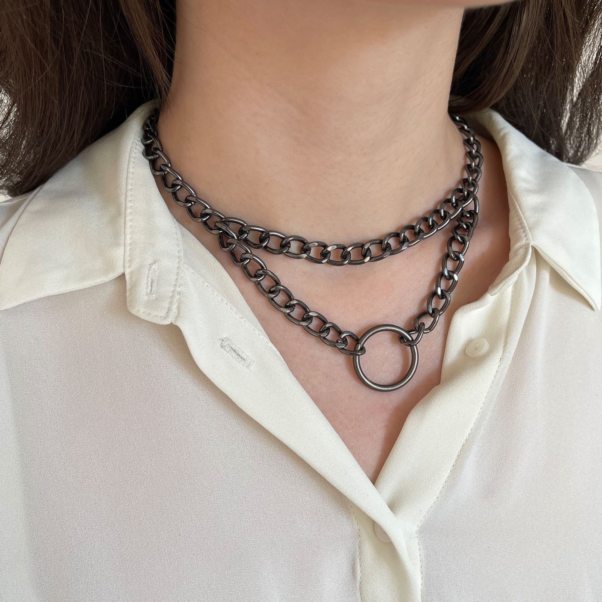 Double chain choker in dark nickel color worn close to the neck, semi-side view. Two metal chains form a layered structure and connect to a round front ring. The choker is secured at the back with a lobster clasp.