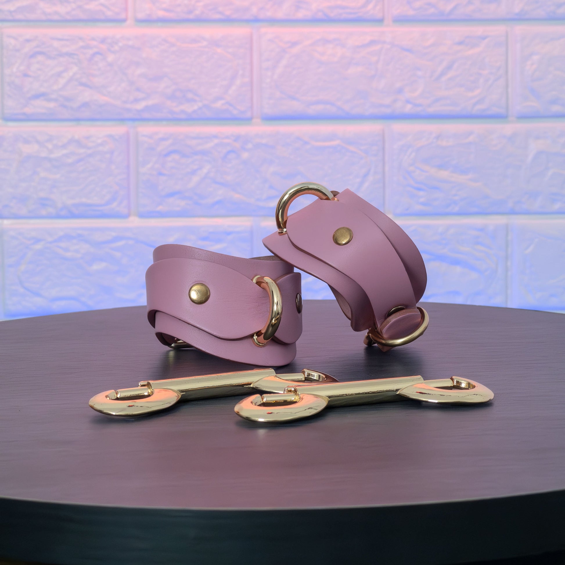 Pink leather handcuffs with gold hardware on a dark surface against a textured light blue background