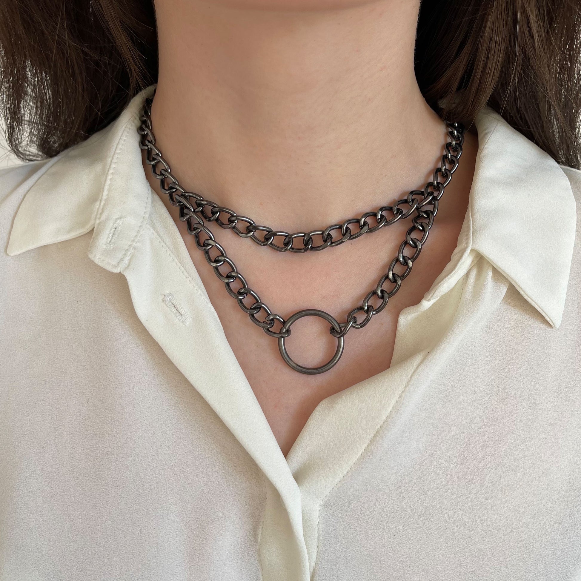 Front-facing image of a gunmetal chain choker with two layers and a central ring. Worn over a white blouse, the matte metallic tone highlights the clean industrial design. An ideal piece for streetwear, grunge, or minimalist aesthetics.