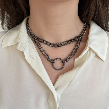 Front-facing image of a gunmetal chain choker with two layers and a central ring. Worn over a white blouse, the matte metallic tone highlights the clean industrial design. An ideal piece for streetwear, grunge, or minimalist aesthetics.
