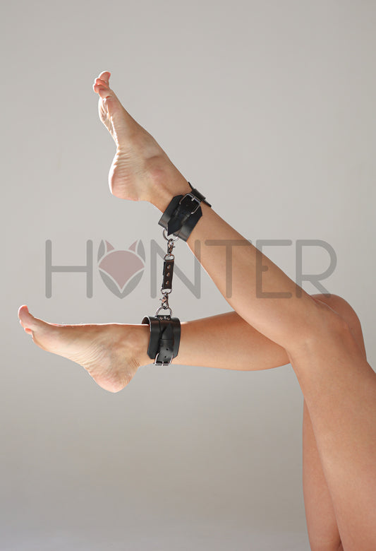 Black genuine leather ankle cuffs worn on a model’s legs, connected with a leather connector using metal carabiners. Each cuff has a 6 cm (2.36 in) wide base, a 2.5 cm (0.98 in) fastening strap with buckle, and a 2.5 cm (0.98 in) metal D-ring, showing how the ankle cuffs fit on the legs and connect together.