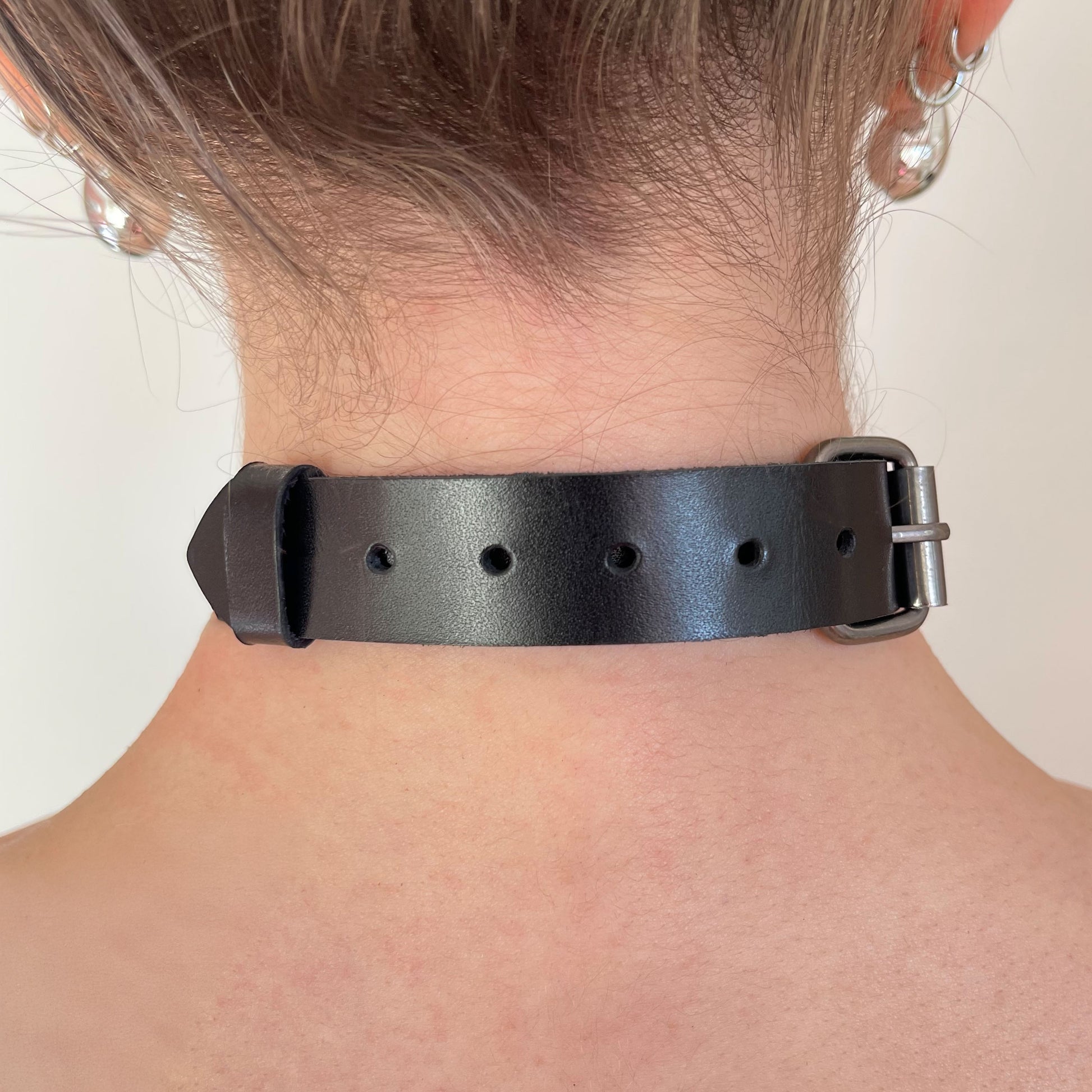 Back view of a black leather choker showing a dark nickel buckle, adjustment holes along the strap, and the strap end secured with a keeper. The 20 mm wide leather strap sits flat against the neck and has a straight cut edge without edge finishing.