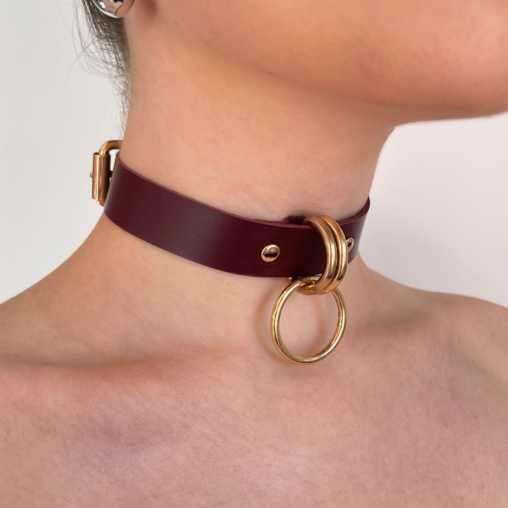 Marsala leather choker worn on the neck in a semi-profile view, gold-tone D-rings holding a round O-ring at the front. The 20 mm wide leather strap with a straight cut edge sits close to the neck, with polished gold hardware as the main visual accent.
