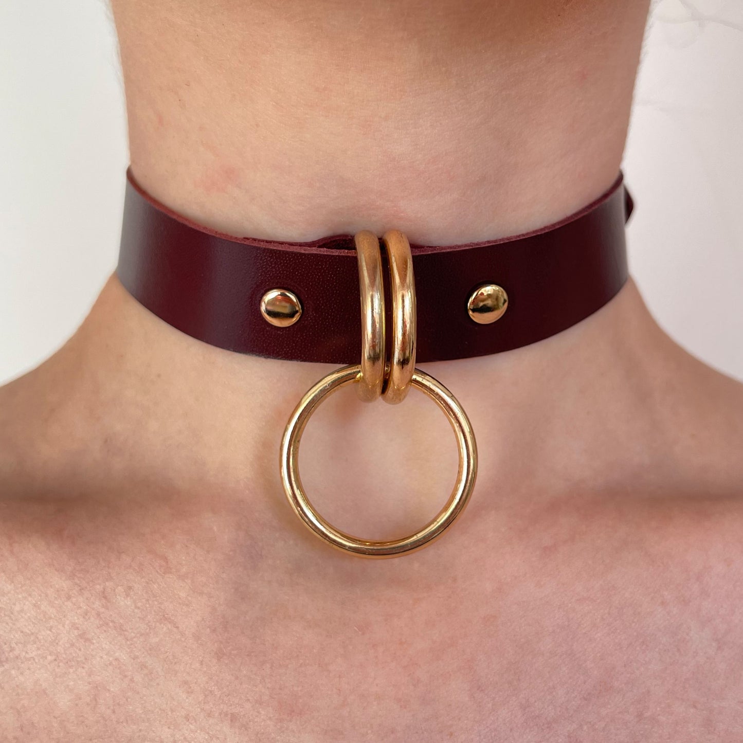 Front view of a marsala leather choker with two gold-tone D-rings holding a round O-ring at the center. The leather strap is evenly aligned around the neck, 20 mm wide, with a straight cut edge and smooth leather surface.