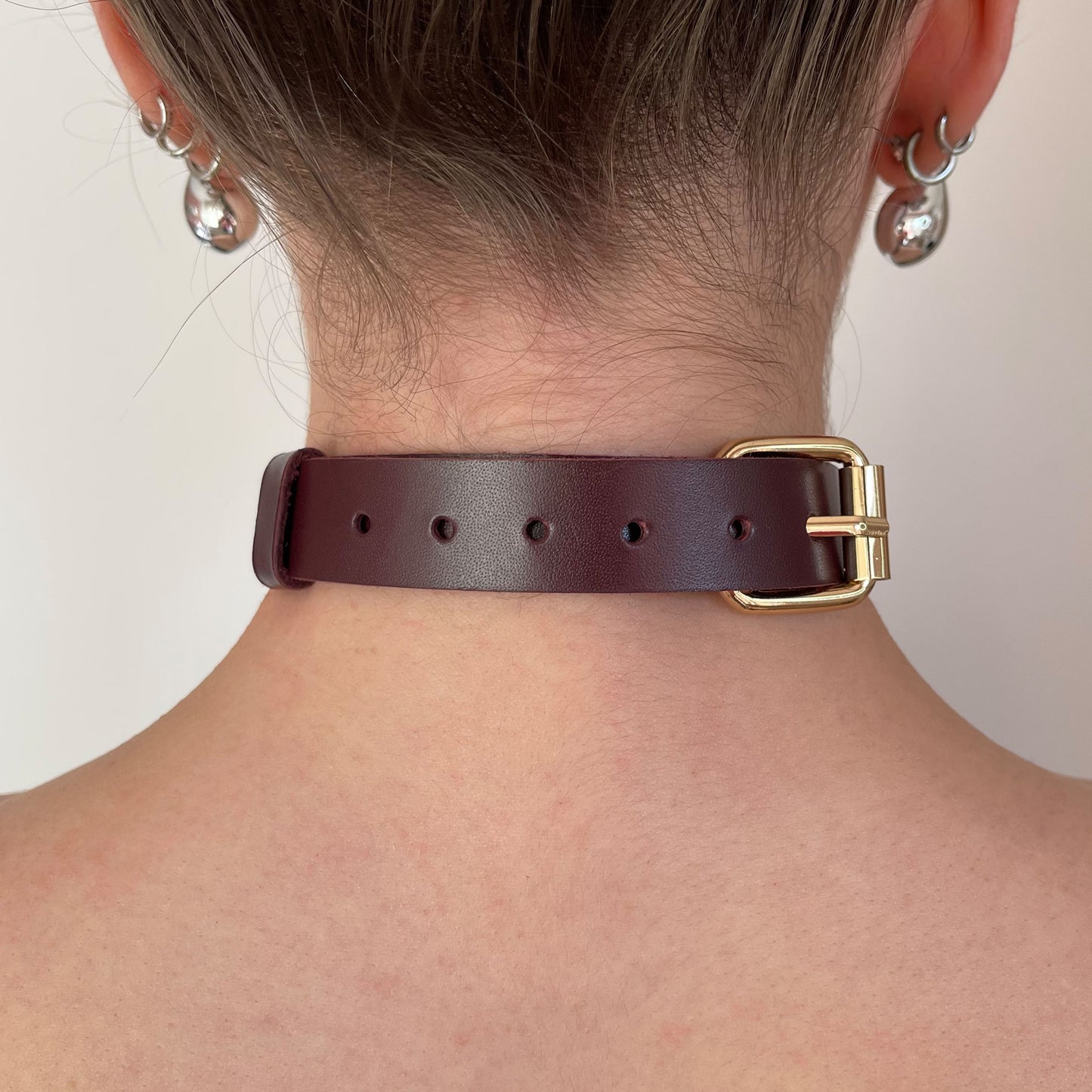 Back view of a marsala leather choker showing a gold-tone buckle, adjustment holes along the strap, and the strap end secured with a keeper. The leather strap lies flat against the neck and has a straight cut edge without edge finishing.