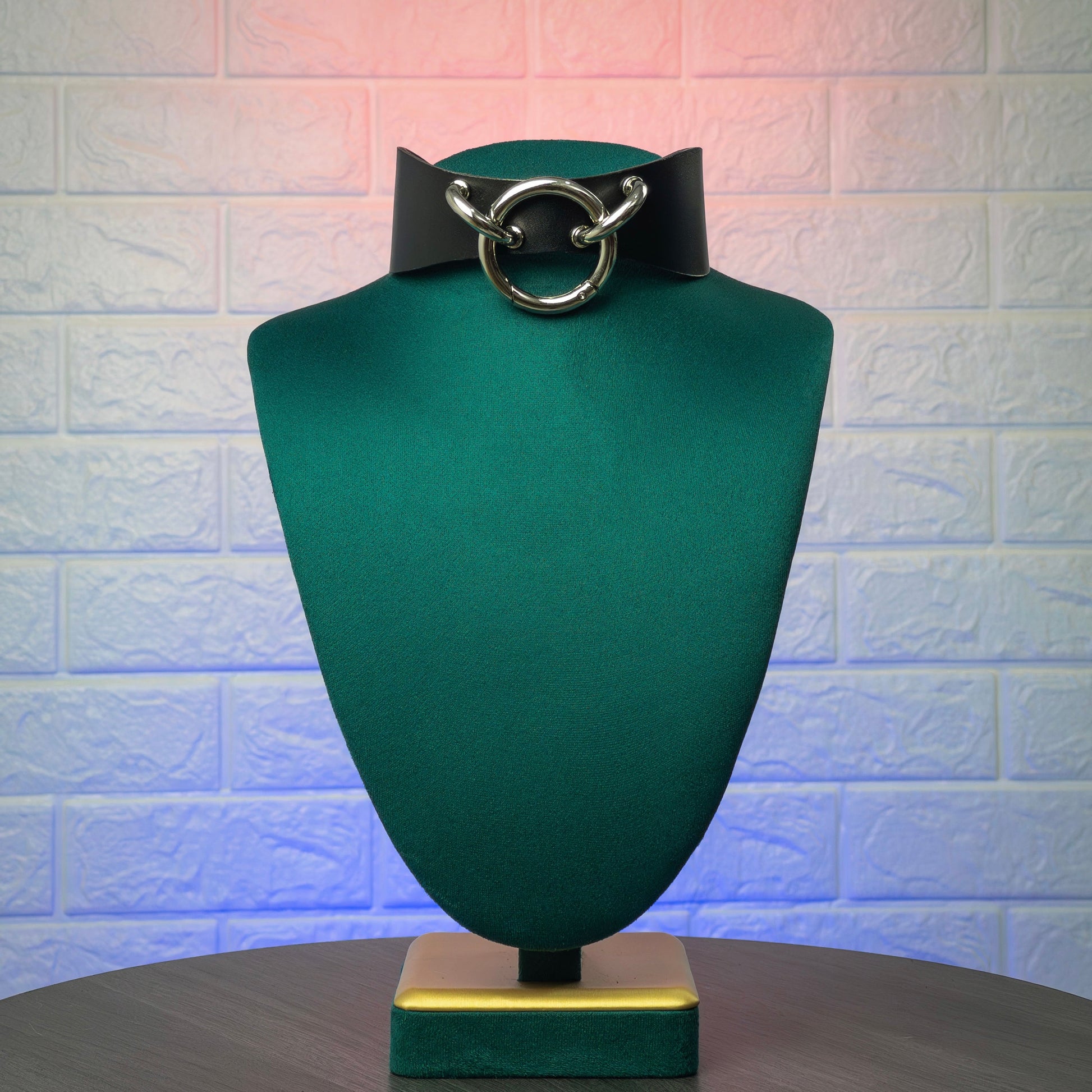 Black leather choker with silver ring on a green mannequin bust against a textured white brick wall.