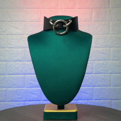 Black leather choker with silver ring on a green mannequin bust against a textured white brick wall.