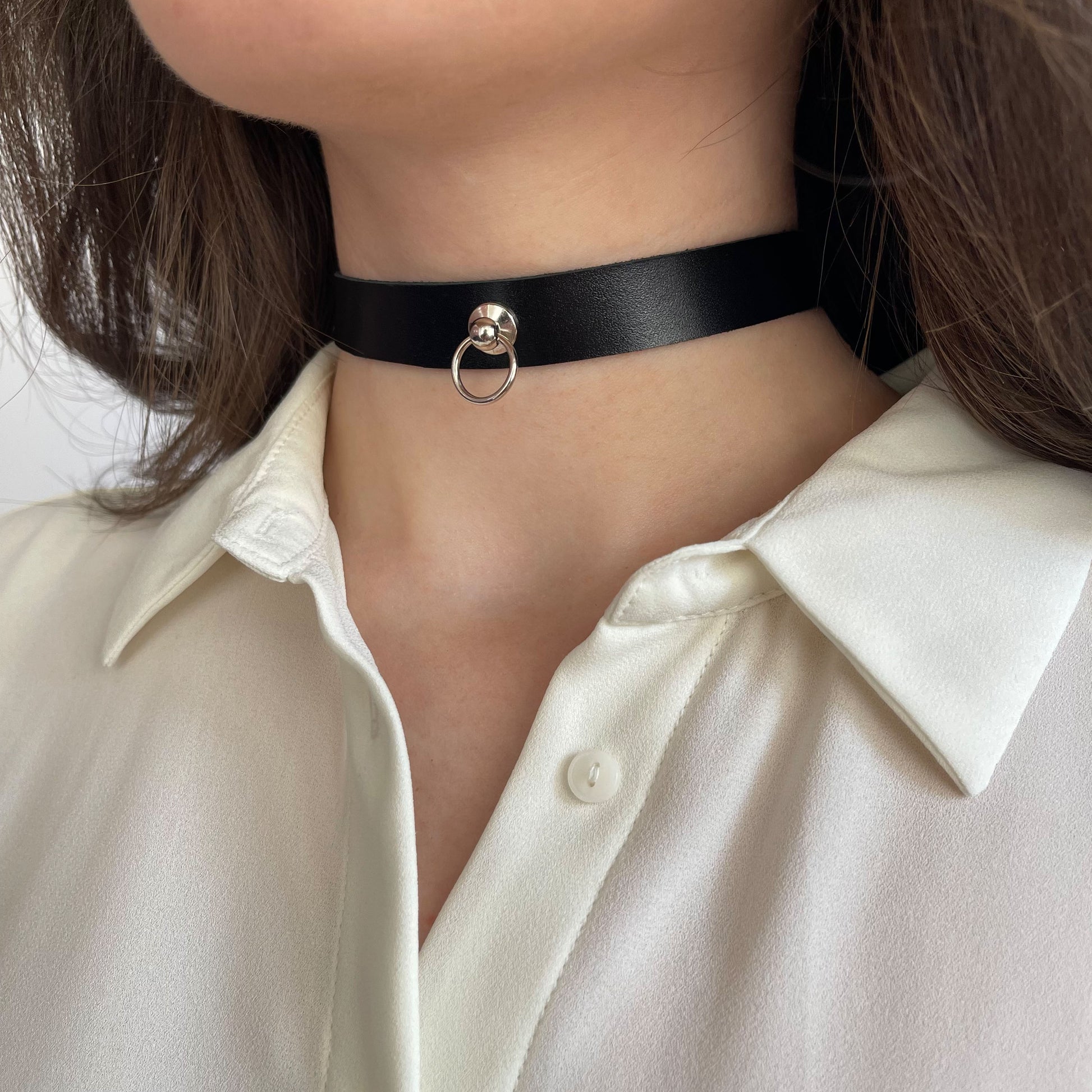 Minimalist black leather choker with a central silver-tone ring. Made of genuine semi-matte leather (1.4–1.8 mm thick), 1.5 cm (0.59 in) wide. Fastened with a buckle. The photo shows a woman’s neck in a white shirt from the side, emphasizing the sleek, structured look of the accessory.