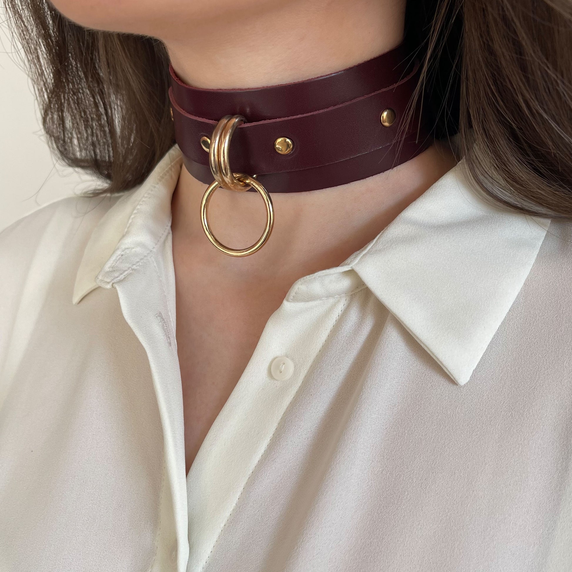 ide leather choker in marsala color with gold hardware worn on the neck in a semi-profile view. The 40 mm wide leather band sits high on the neck, with two 20 mm D-rings holding a 25 mm round O-ring at the front. The 20 mm strap and buckle are partially visible.