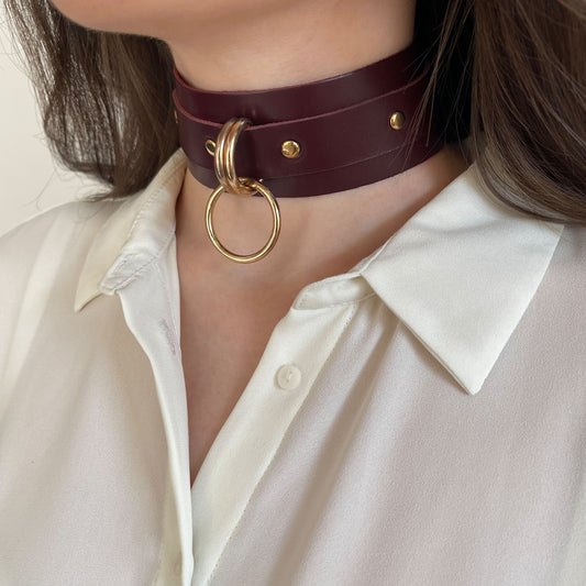 ide leather choker in marsala color with gold hardware worn on the neck in a semi-profile view. The 40 mm wide leather band sits high on the neck, with two 20 mm D-rings holding a 25 mm round O-ring at the front. The 20 mm strap and buckle are partially visible.