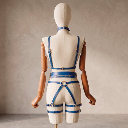 A blue leather harness-style outfit with matching belt and choker displayed on a cream mannequin with wooden arms, photographed from the back against a textured beige studio wall.