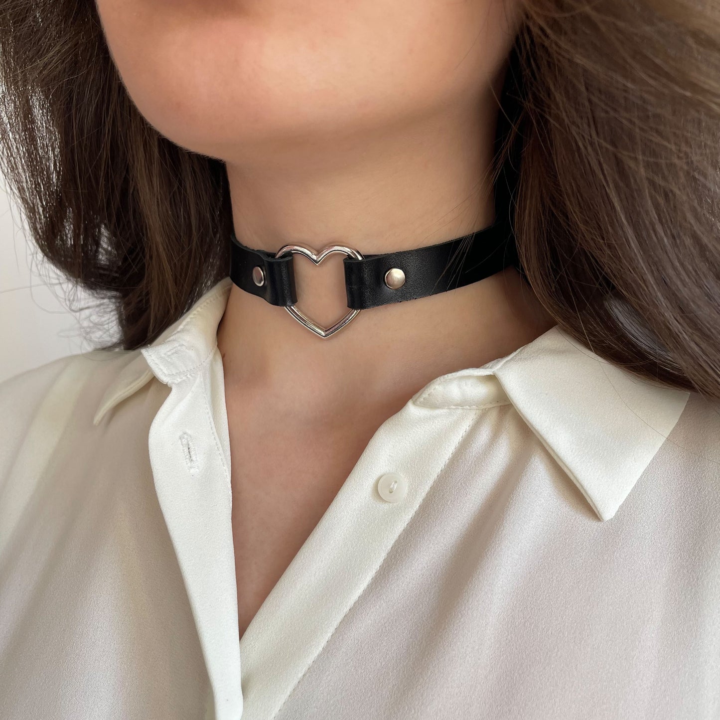 Black leather choker worn on the neck, shown from a semi-side angle. The strap is 15 mm (0.59 in) wide, made of smooth natural leather with moderate firmness. At the center is a silver-tone metal heart-shaped ring, 30 mm (1.18 in) in diameter, fixed with silver rivets. The choker sits flat and follows the neck line.