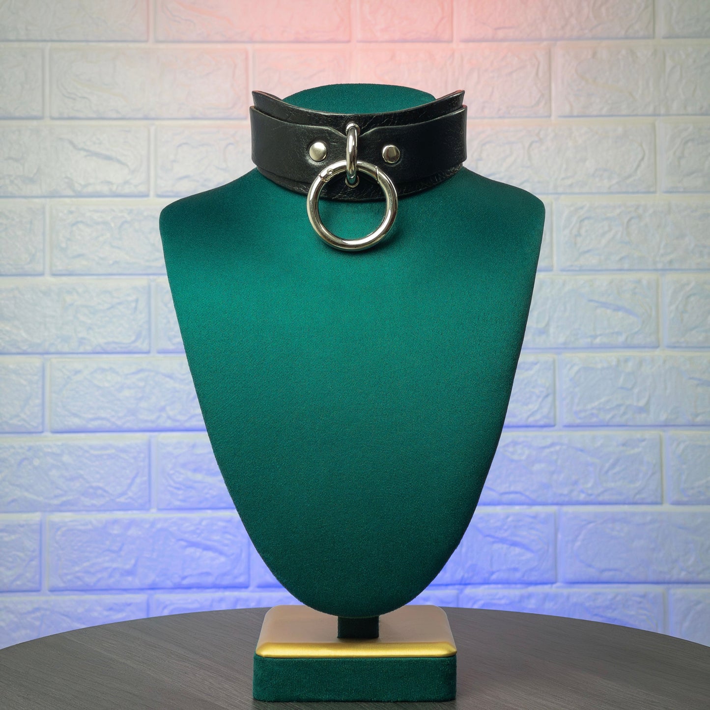 Black leather choker with a large ring on a green mannequin bust against a textured white wall.