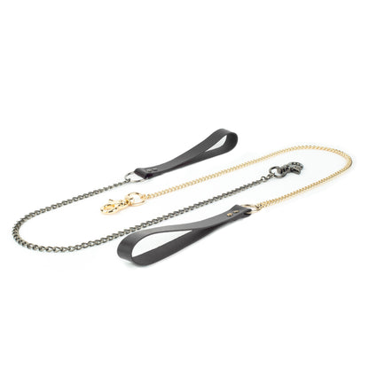 BDSM leather chain leash on a white background, black leather hand loop connected to a 60 cm metal chain with a carabiner fixed directly to the chain end. The photo shows the same leash with two hardware finishes, dark nickel and gold, laid flat for comparison.