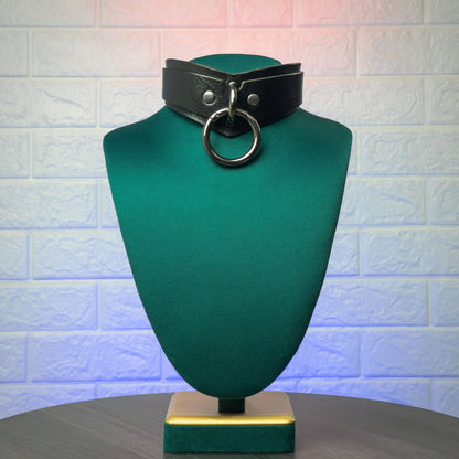 Black leather choker with a large ring on a green mannequin bust against a textured white wall.