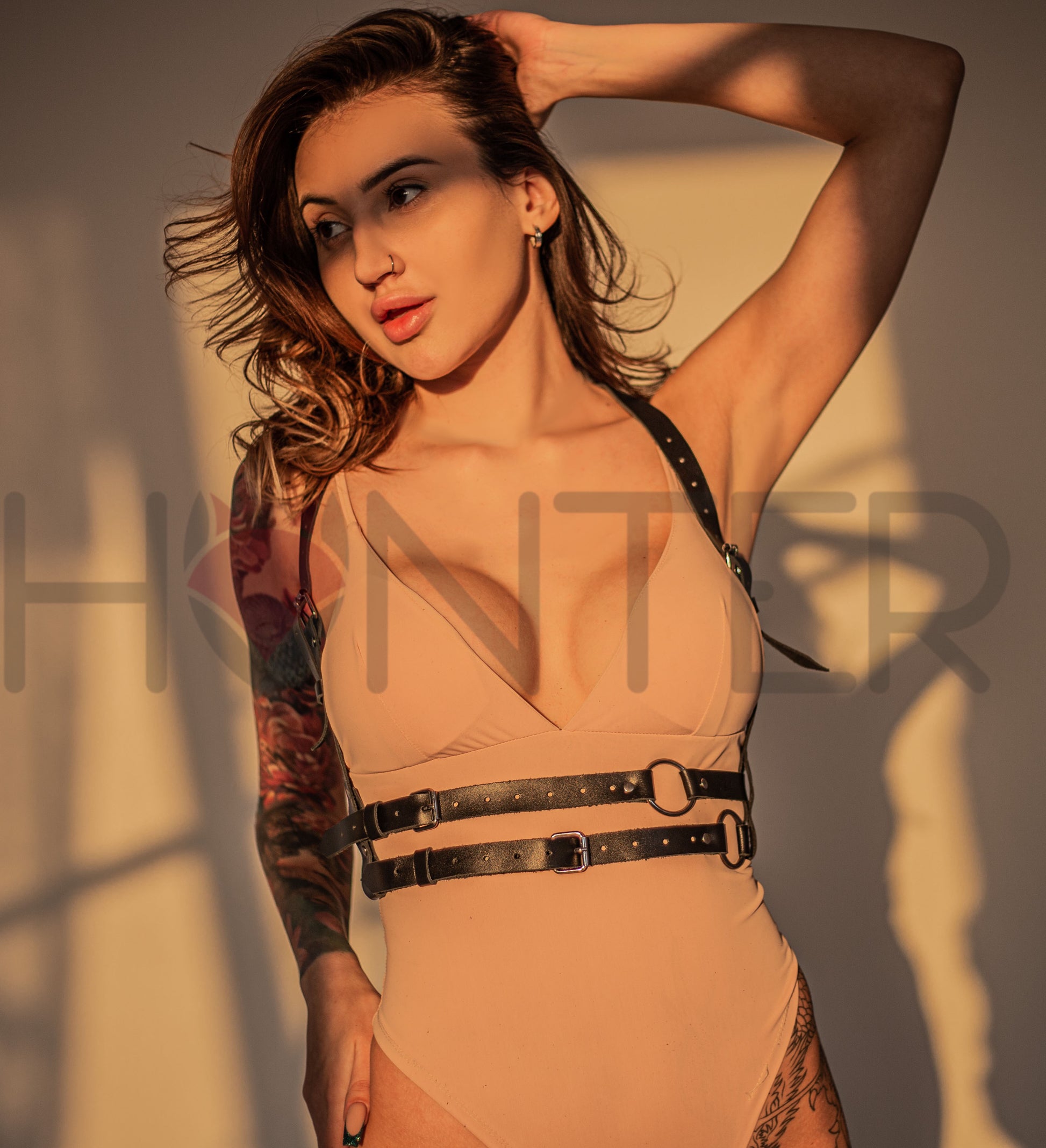 Front angled view of a black leather harness with a double waist construction and ring details on a model in a nude bodysuit. The image shows the upper and lower waist straps, the front rings, the shoulder straps descending along the torso, and the fitted shape of the harness in warm directional light.