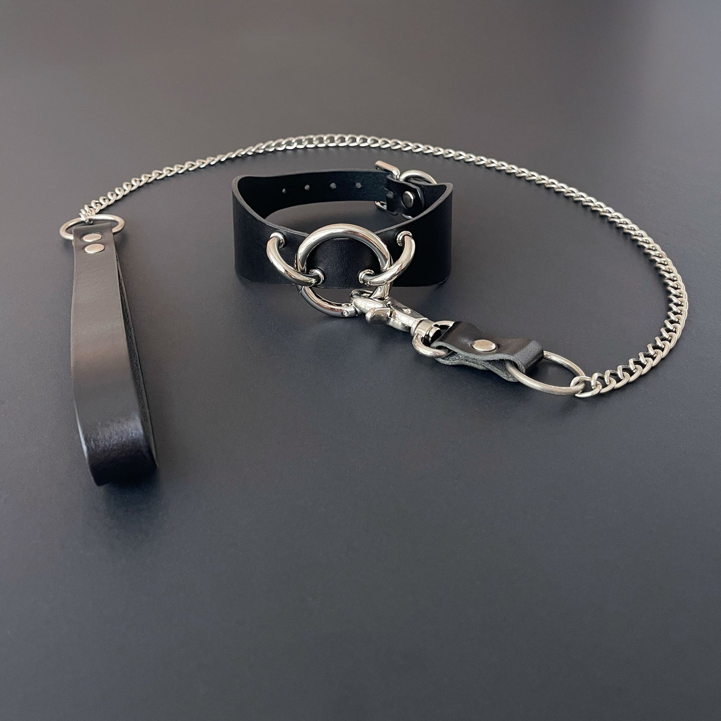 Black leather collar with silver chain and strap on a dark surface