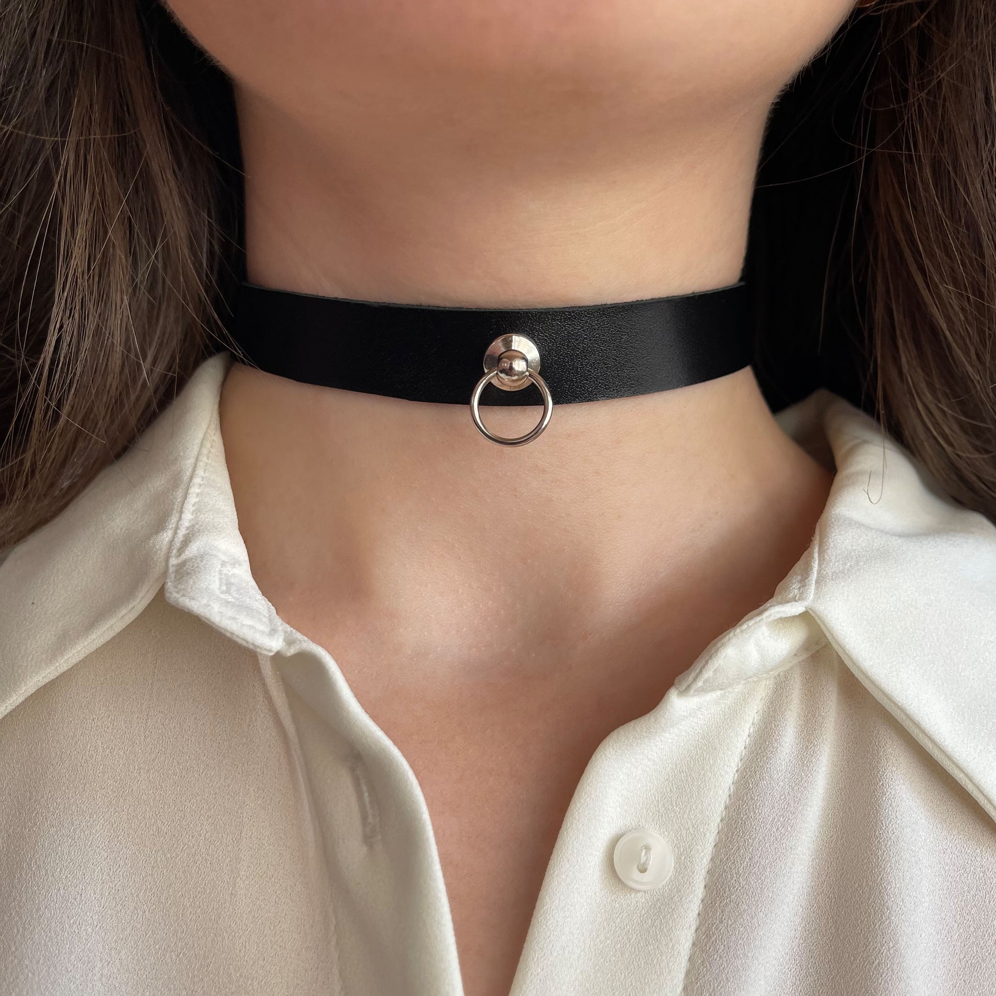 Classic black leather choker with a silver ring detail at the center. Crafted from genuine semi-matte leather, 1.5 cm (0.59 in) wide and 1.4–1.8 mm thick. Buckle closure. Close-up of a woman’s neck in a white shirt, highlighting the contrast and bold simplicity of the piece.