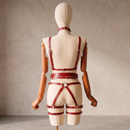 A red leather harness-style accessory with straps, buckles, and a matching waist belt is displayed on a beige mannequin with wooden arms, photographed against a neutral textured wall.